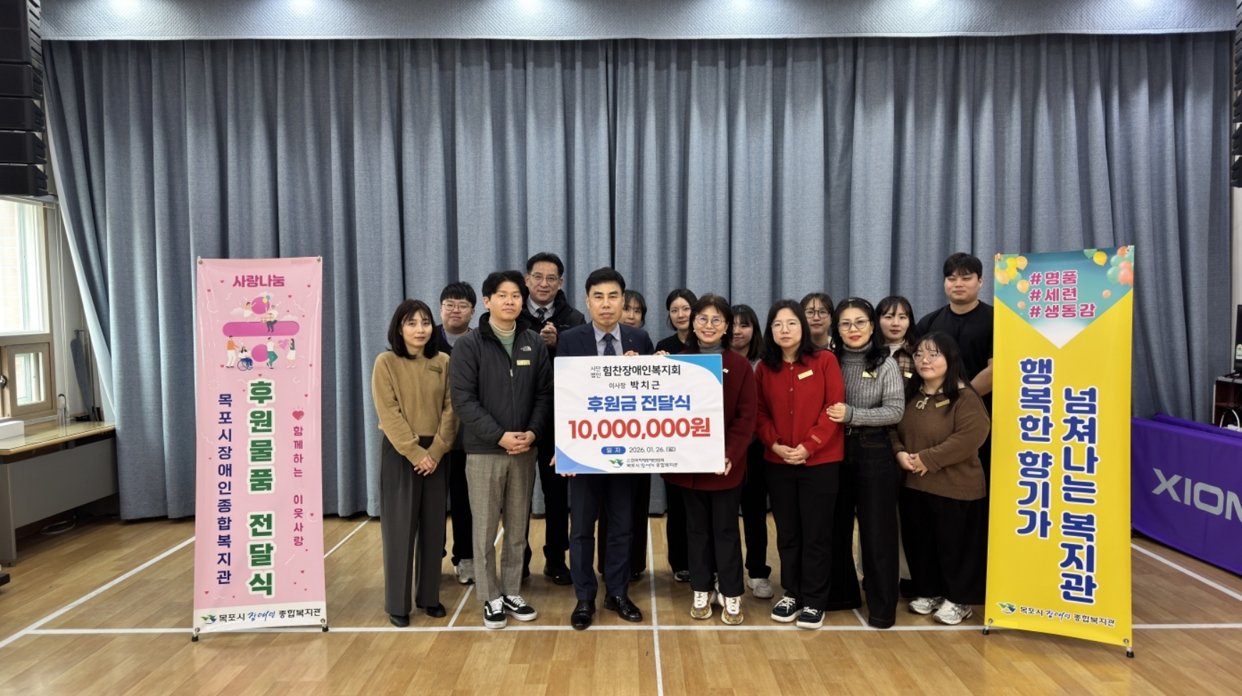 사단법인 힘찬장애인복지회 박치근 이사장이 목포시장애인종합복지관에 후원금 1,000만 원을 전달하고 기념촬영을 하고 있다. 실내 강당 무대 위에서 박치근 이사장과 복지관 관계자가 함께 '사단법인 힘찬장애인복지회 이사장 박치근 후원금 전달식 10,000,000원'이라고 적힌 피켓을 들고 서 있다. 그 뒤로 10여 명의 관계자들이 함께 서 있으며, 사진 좌측에는 '사랑나눔 후원 물품 전달식'이라고 적힌 분홍색 배너가, 우측에는 '#명품 #세련 #생동감 넘쳐나는 복지관 행복한 향기가'라고 적힌 노란색 배너가 세워져 있다.